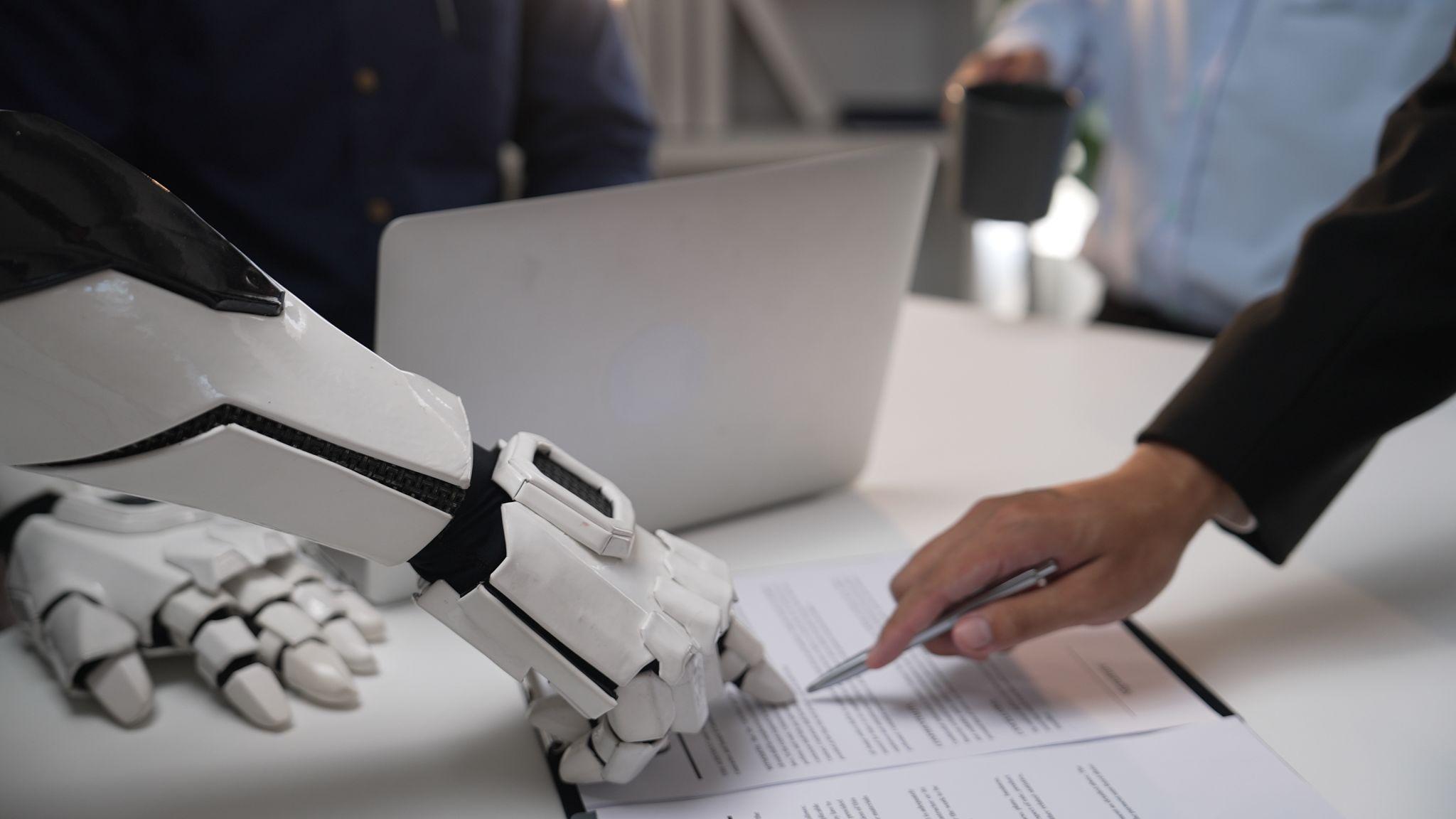 Robot arm and human hand reviewing a document together at a desk — AI and human collaboration in practice
